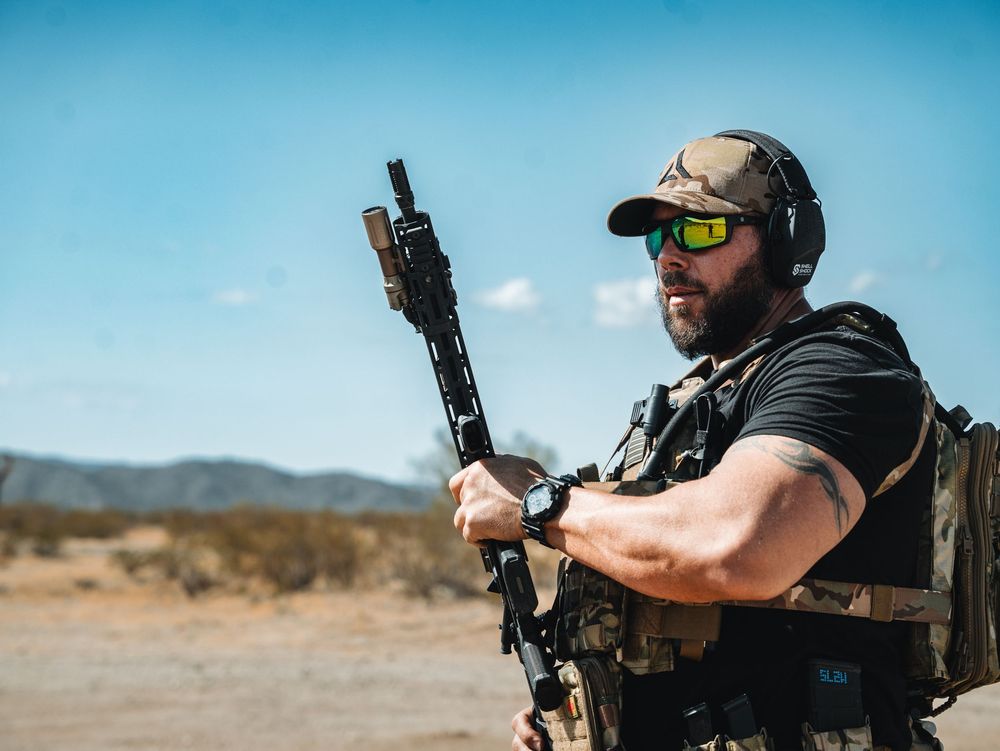 Tactical shooter wearing protective gear and holding rifle stands in desert landscape against blue sky.