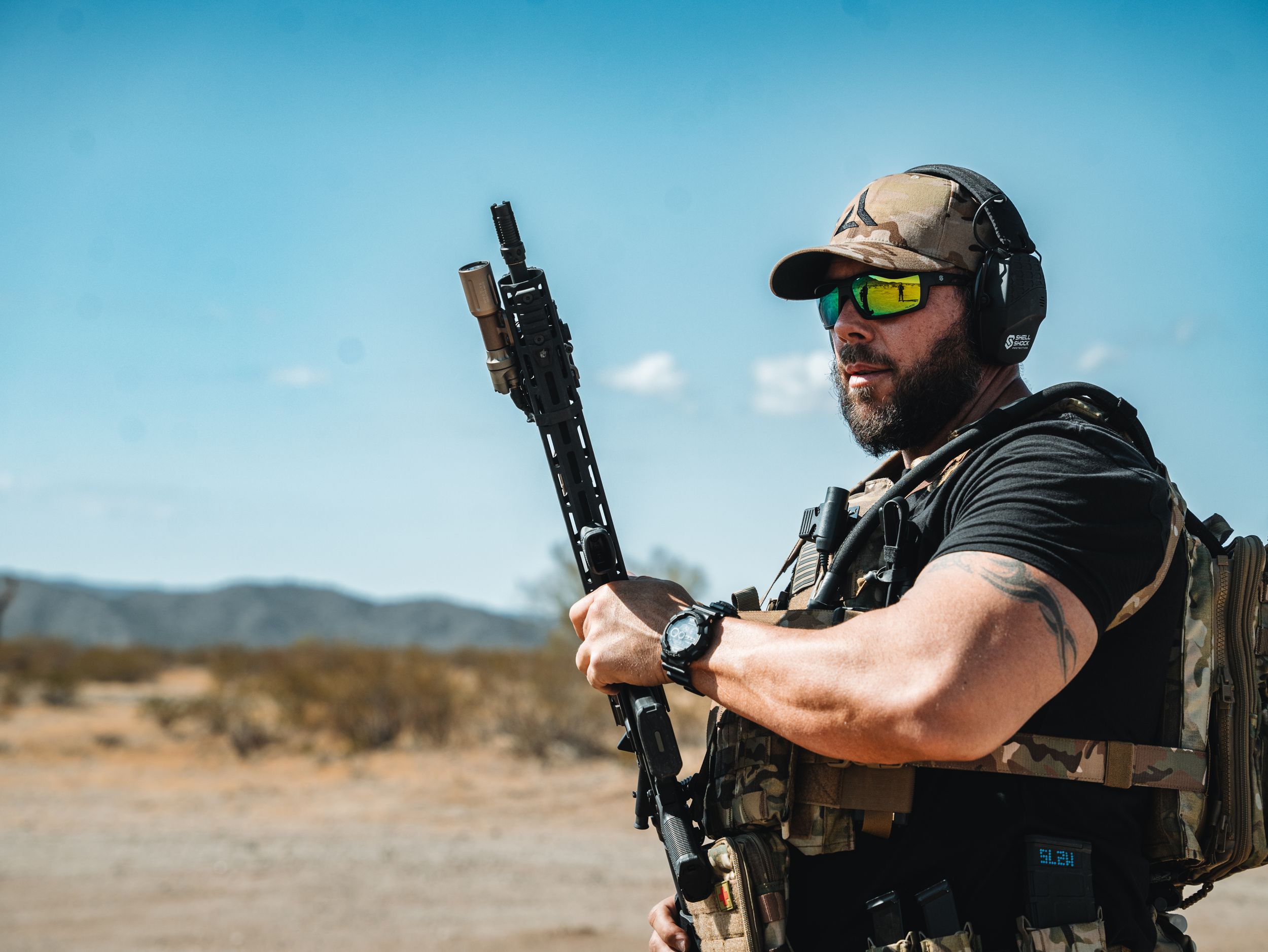 Tactical shooter wearing protective gear and holding rifle stands in desert landscape against blue sky.