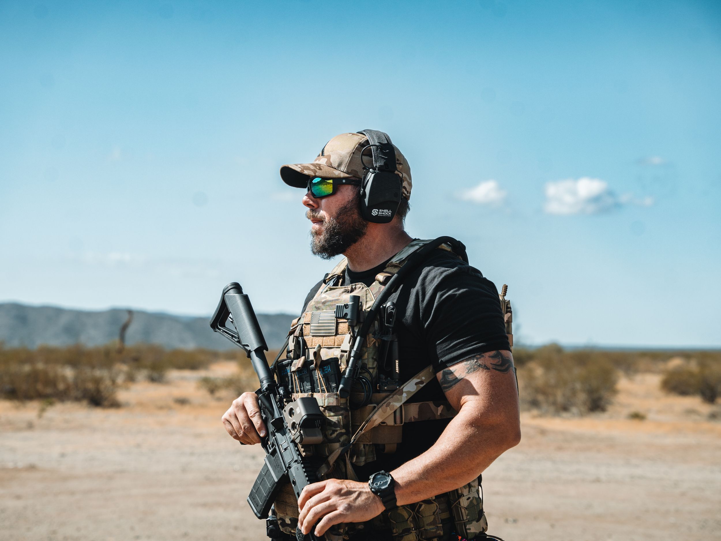 A person wearing tactical gear aims a rifle in a desert environment during shooting practice.