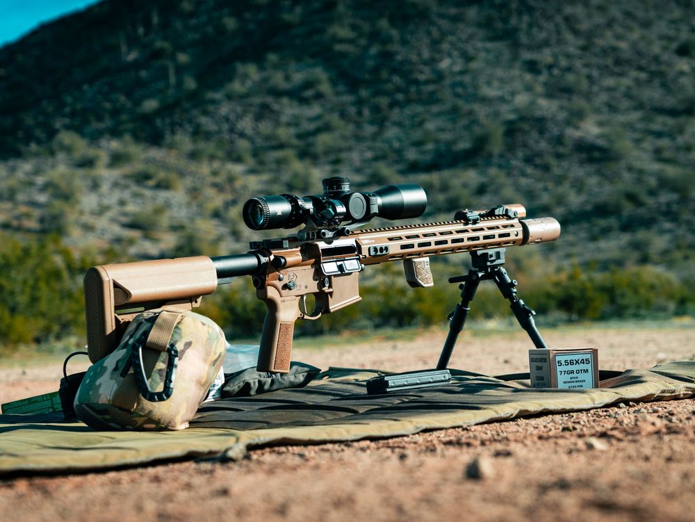 Long-range rifle with scope and bipod set up on shooting mat in desert terrain.