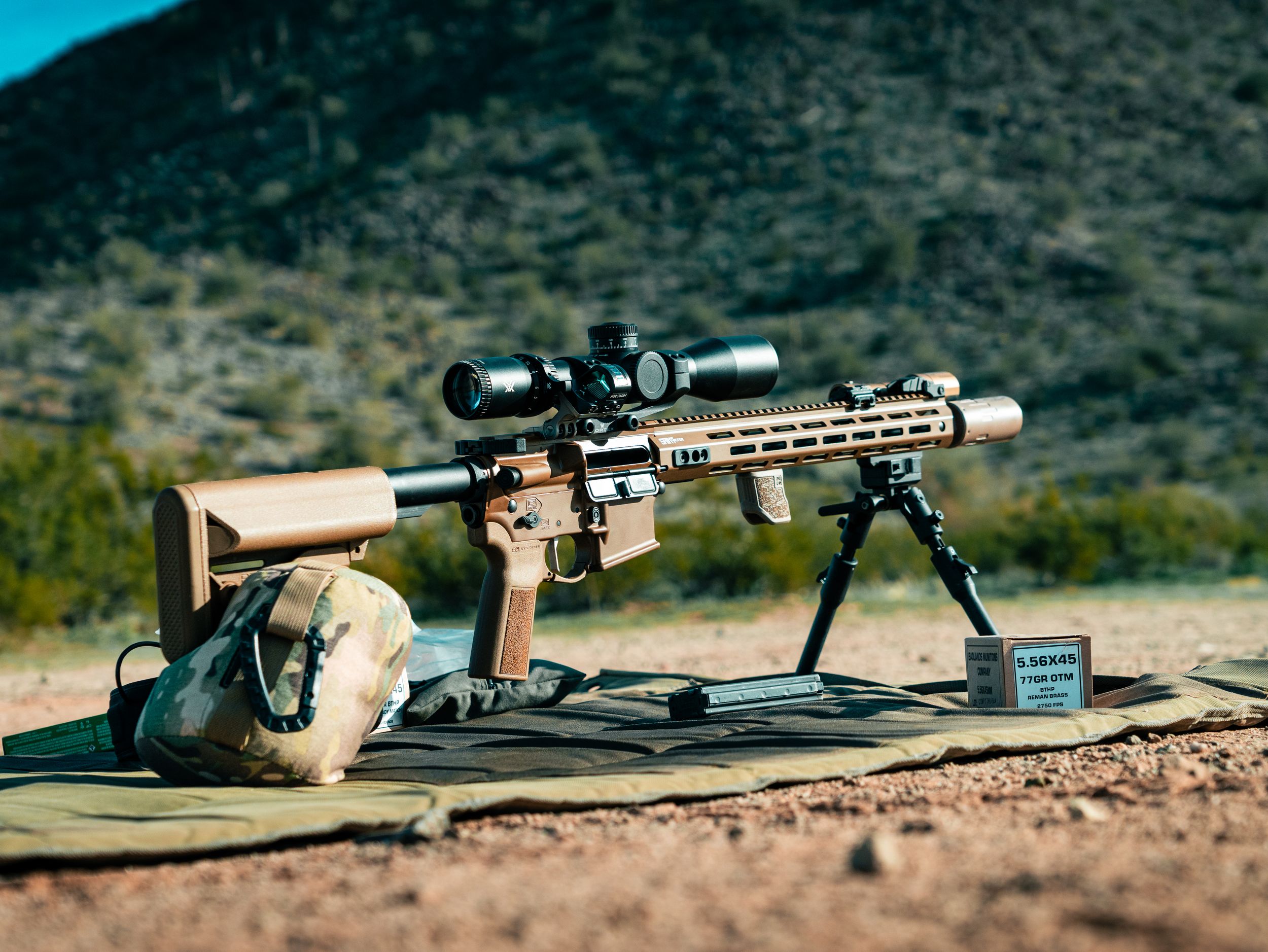 Long-range rifle with scope and bipod set up on shooting mat in desert terrain.