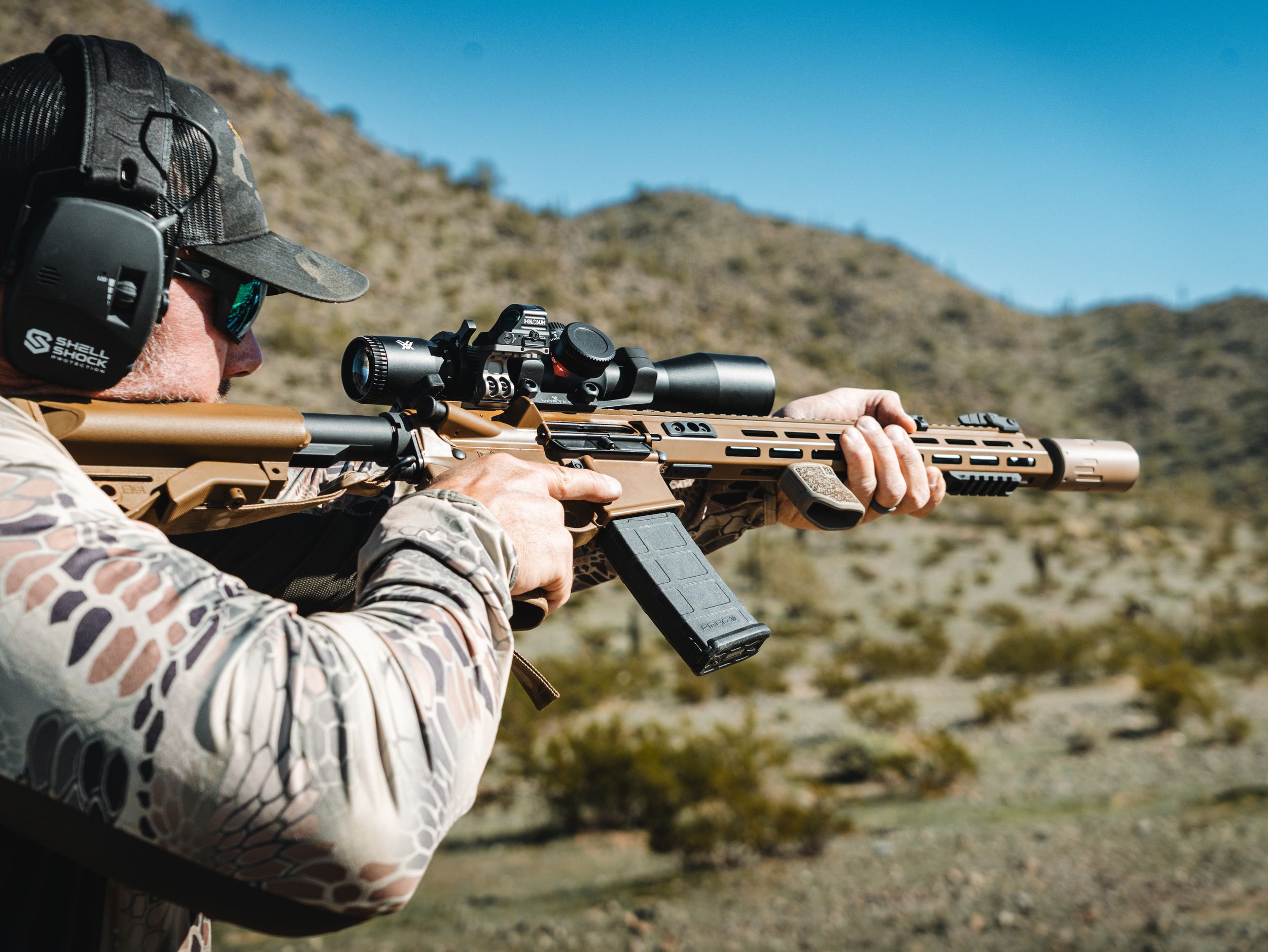 Shooter in desert camouflage aiming tactical rifle against mountain backdrop.