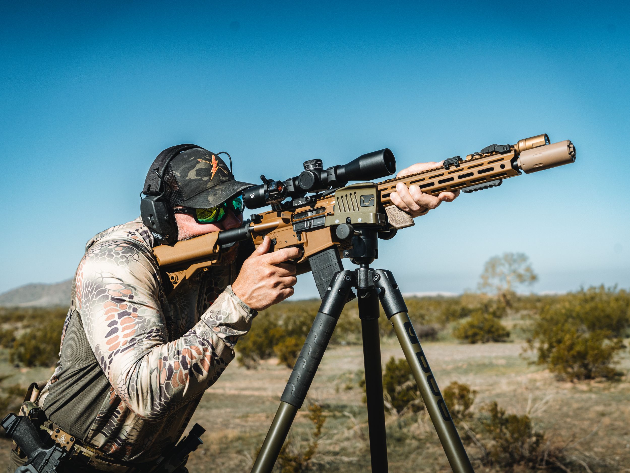 A shooter aims a long-range rifle with a scope mounted on a tripod in an outdoor desert setting.
