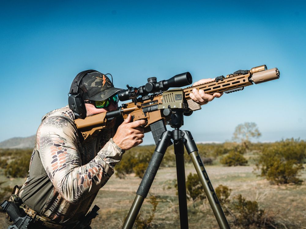 A shooter aims a long-range rifle with a scope mounted on a tripod in an outdoor desert setting.