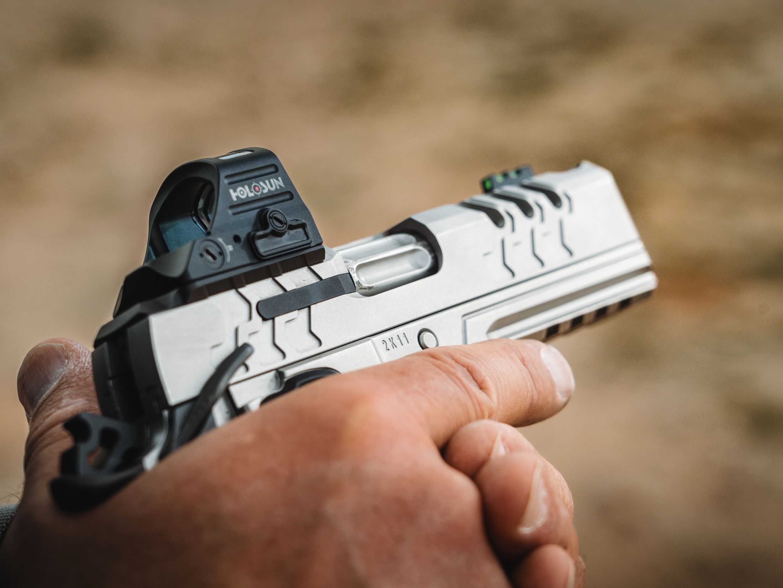 Close-up of hands holding silver pistol with mounted optic against desert background.