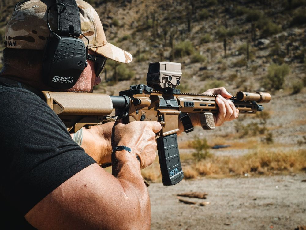 A shooter takes aim with a rifle at an outdoor shooting range in a rocky desert environment.