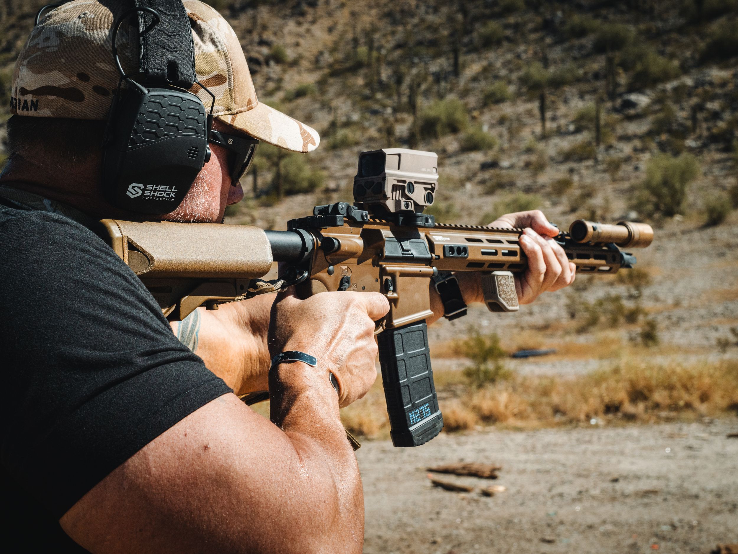 A shooter takes aim with a rifle at an outdoor shooting range in a rocky desert environment.