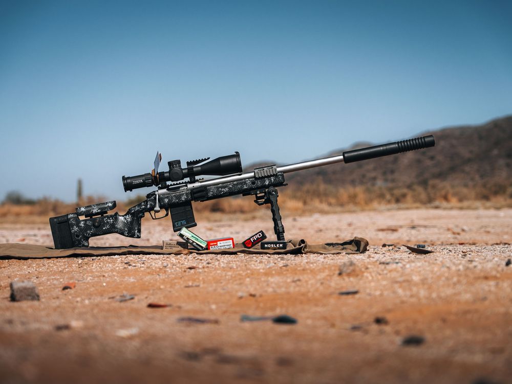 Long range rifle with scope lies on dusty desert ground during target practice shooting session.