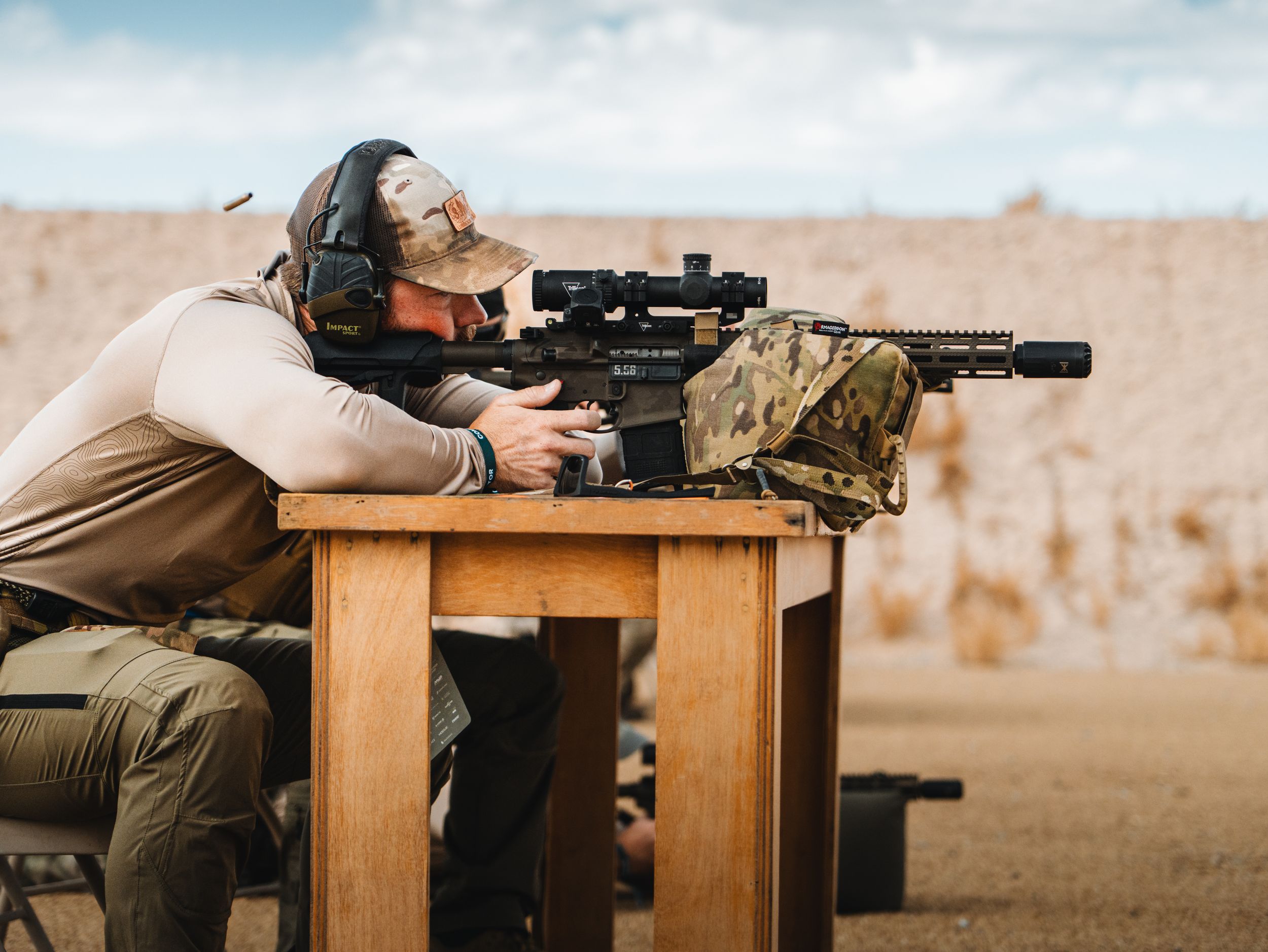 A shooter in tactical gear aims a scoped rifle while positioned at a wooden bench on an outdoor range.