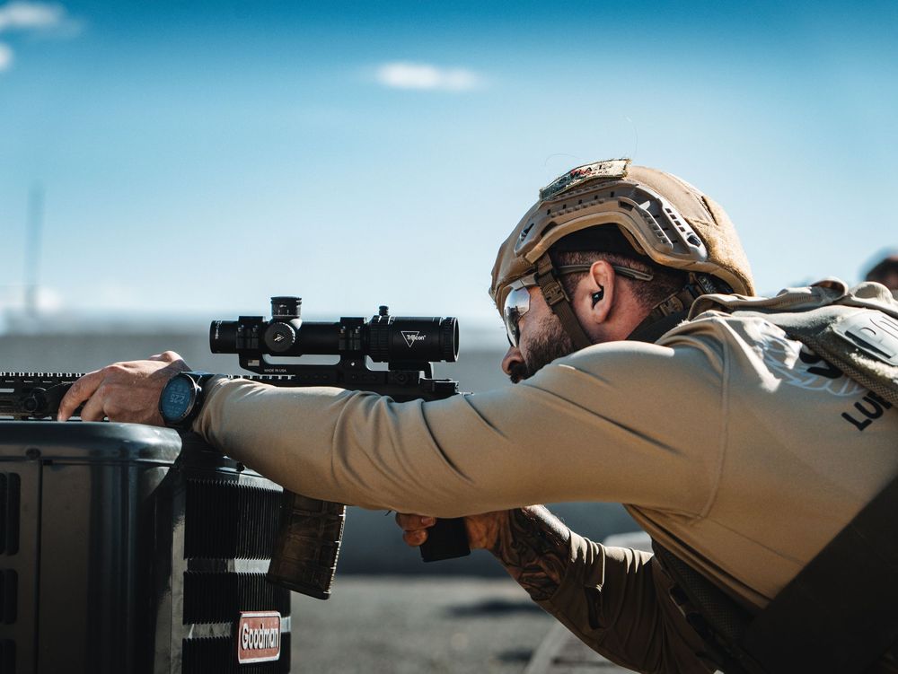 A swat officer taking a shot from behind an ac unit on a roof.