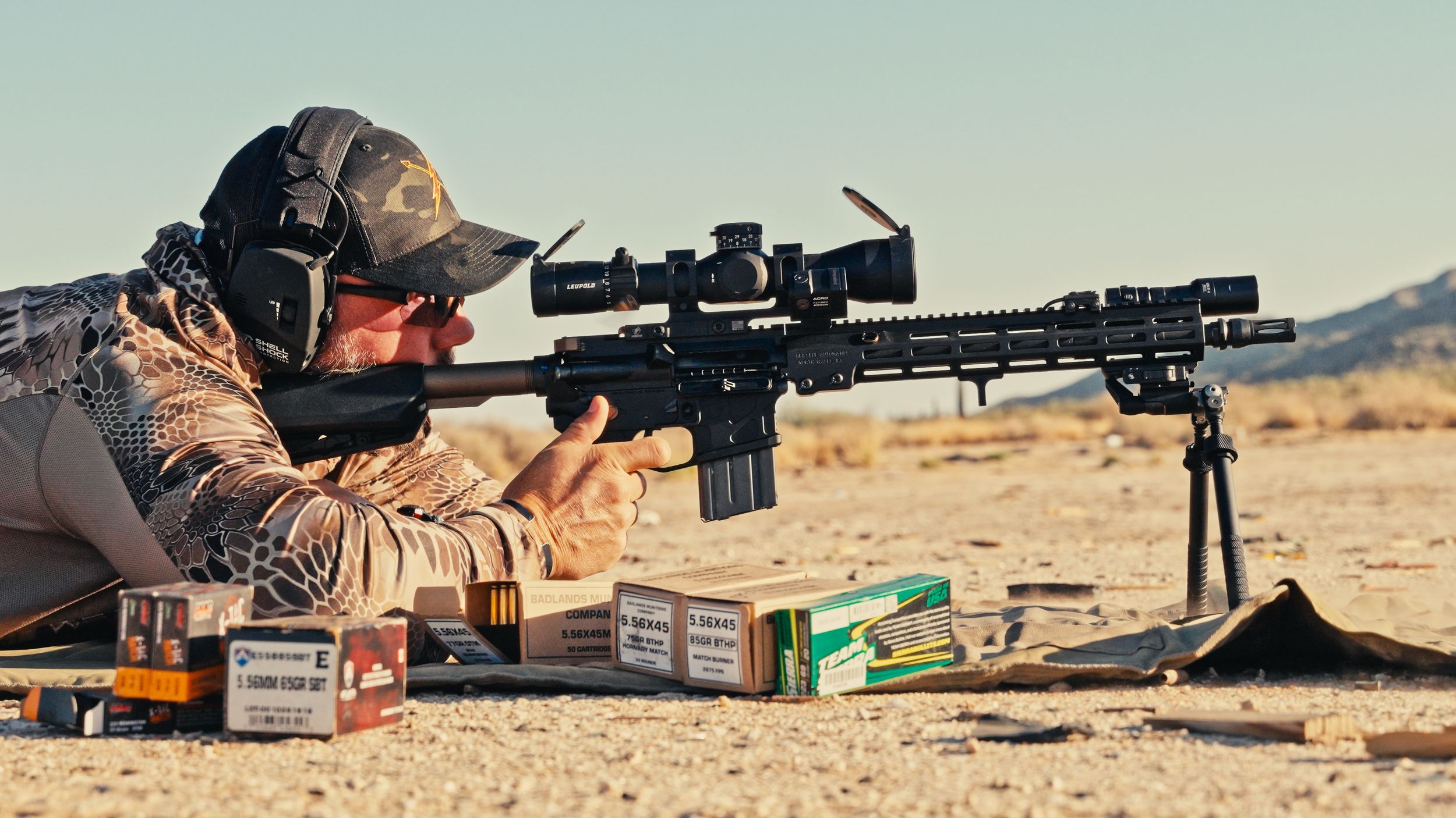 A row of people practicing shooting positions with rifles at an outdoor desert shooting range.