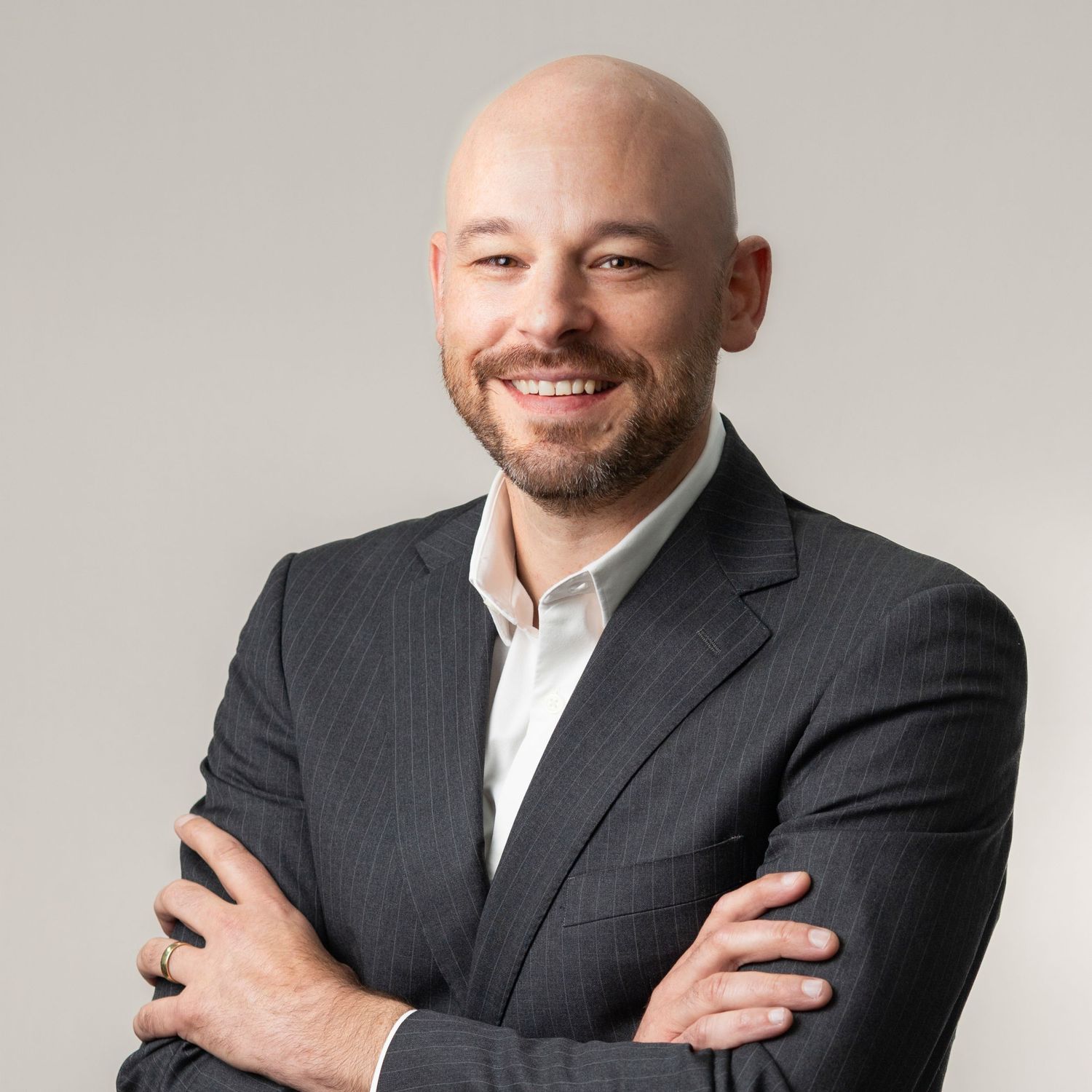Professional headshot of a bald person in a dark pinstripe suit and white shirt smiling with arms crossed.