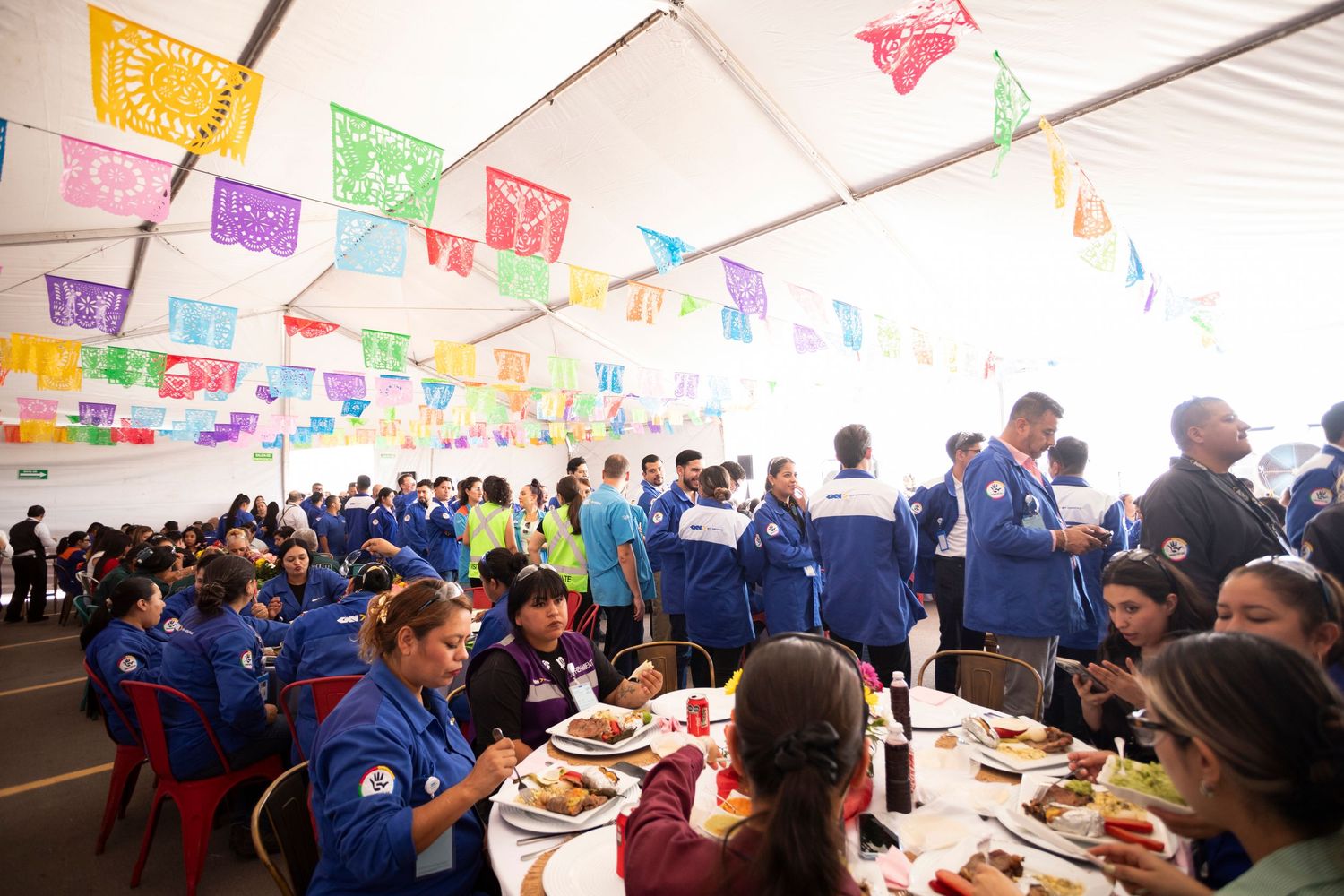 Colorful papel picado banners hang from ceiling at festive gathering with people seated at long tables.