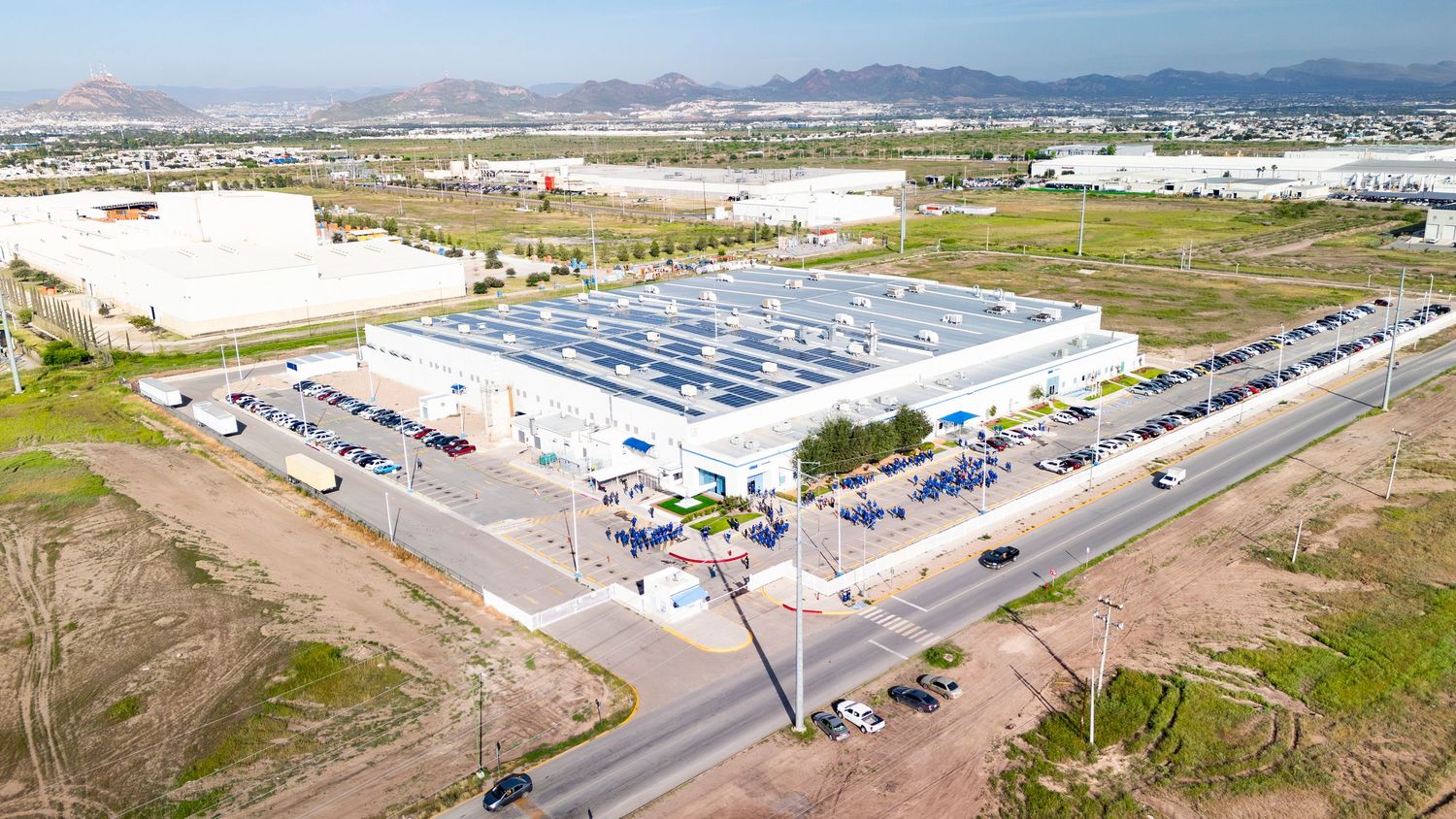 Panoramic aerial view of industrial warehouses and solar panel arrays in an arid desert landscape with mountains in background.