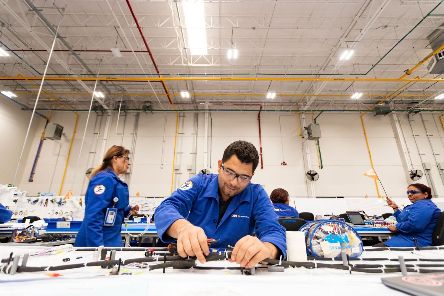Workers in blue uniforms inspect and assemble electronic components at a manufacturing facility workstation.