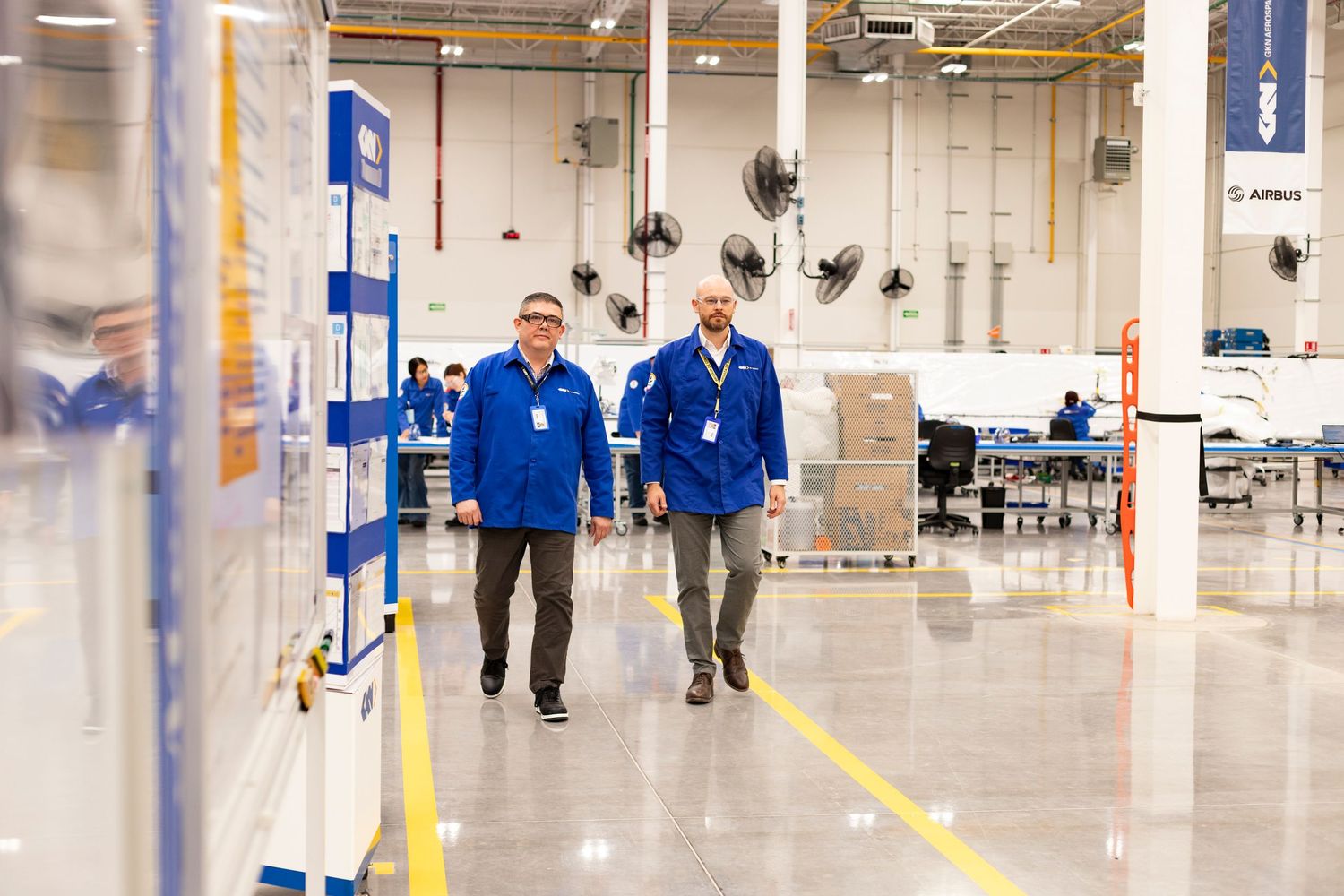 Workers walk through a large industrial warehouse facility with yellow floor markings and blue equipment visible.