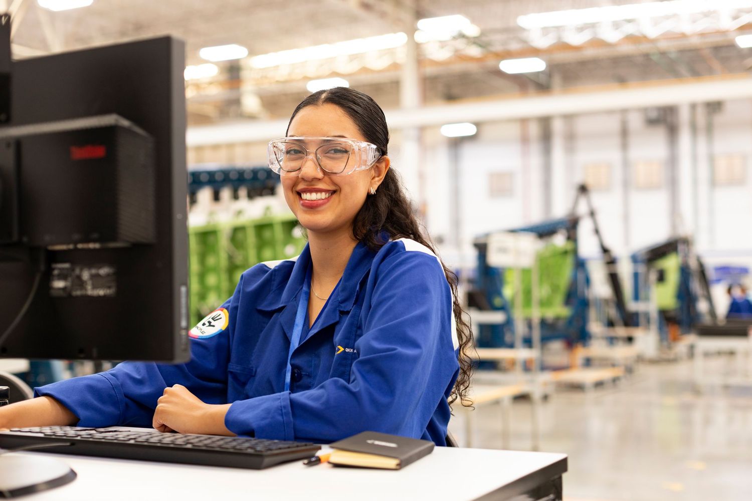 Professional wearing safety glasses and blue lab coat works at manufacturing facility while smiling at computer screen.