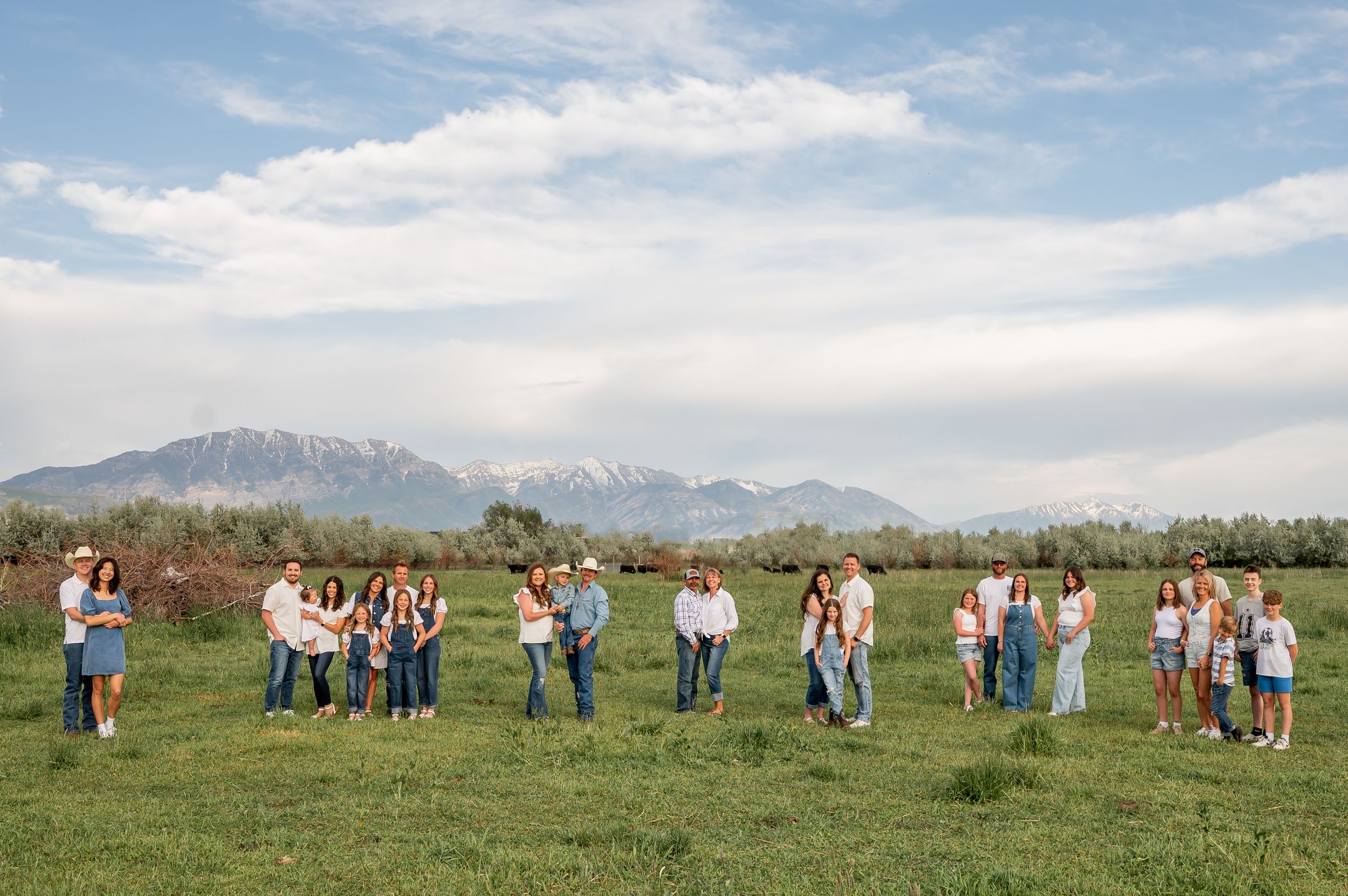 A group of people stand in a grassy field with snow-capped mountains in the background on a sunny day.
