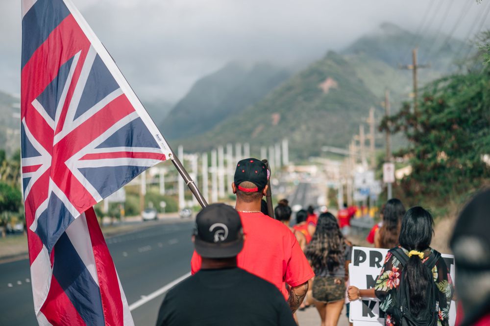 People holding Hawaiian flags gather along a road with mountains in the background.