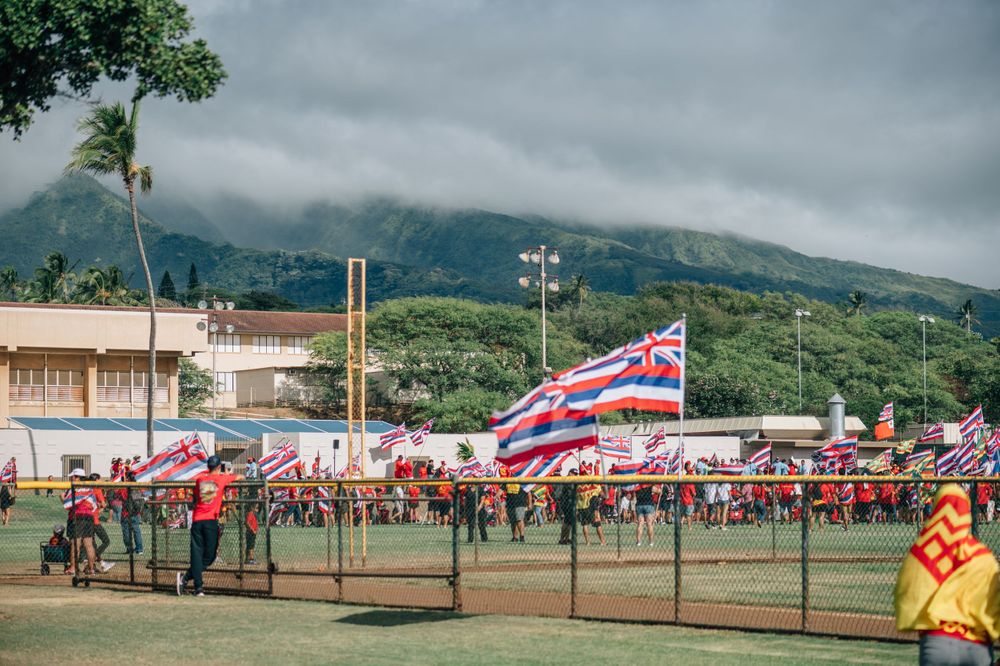 Large gathering on a field with Hawaiian flags and mountains visible in the distance.