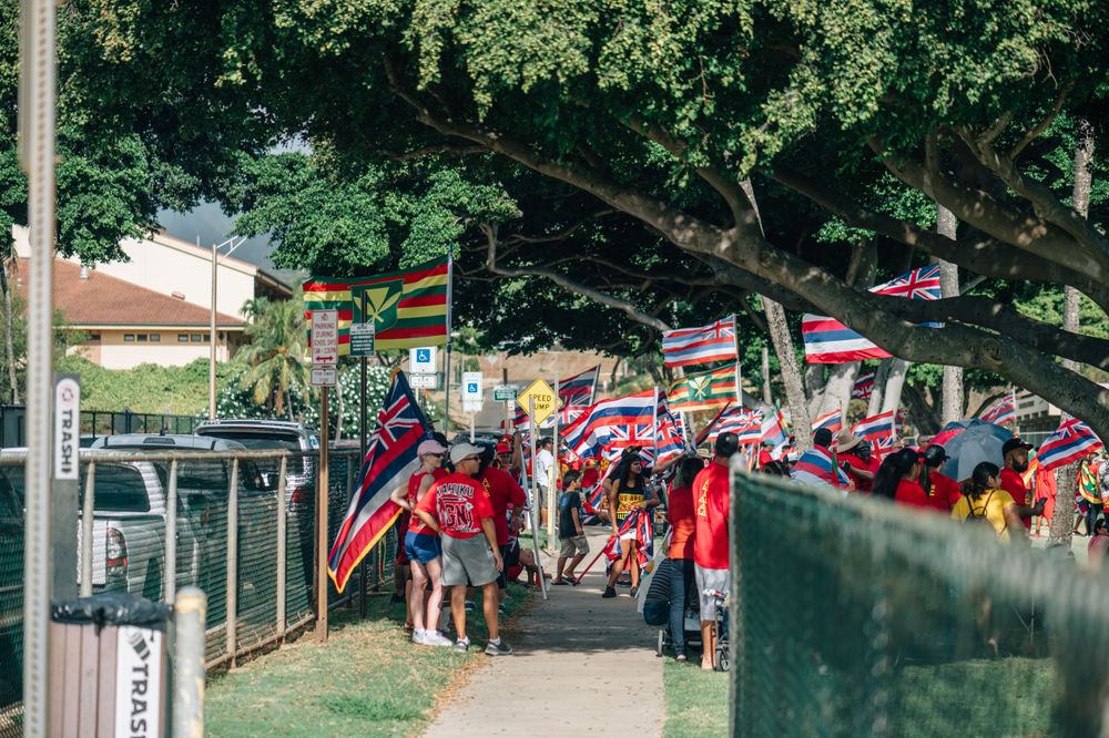 People gather under trees waving Hawaiian flags at an outdoor event.