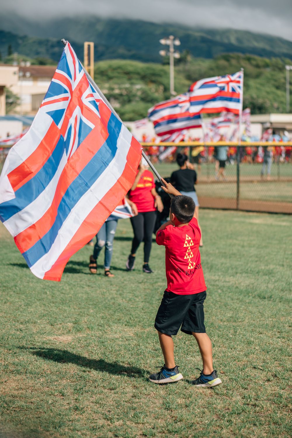 Children wave Hawaiian state flags on a grassy field with mountains behind them.