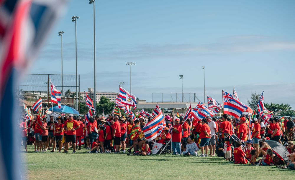 Crowd in red shirts stands together waving Hawaiian flags against blue sky.
