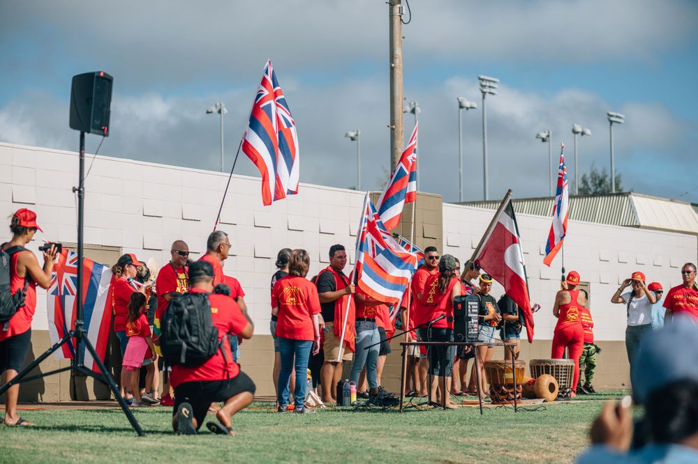 Crowd of supporters in red shirts waving Hawaiian flags at a political gathering.
