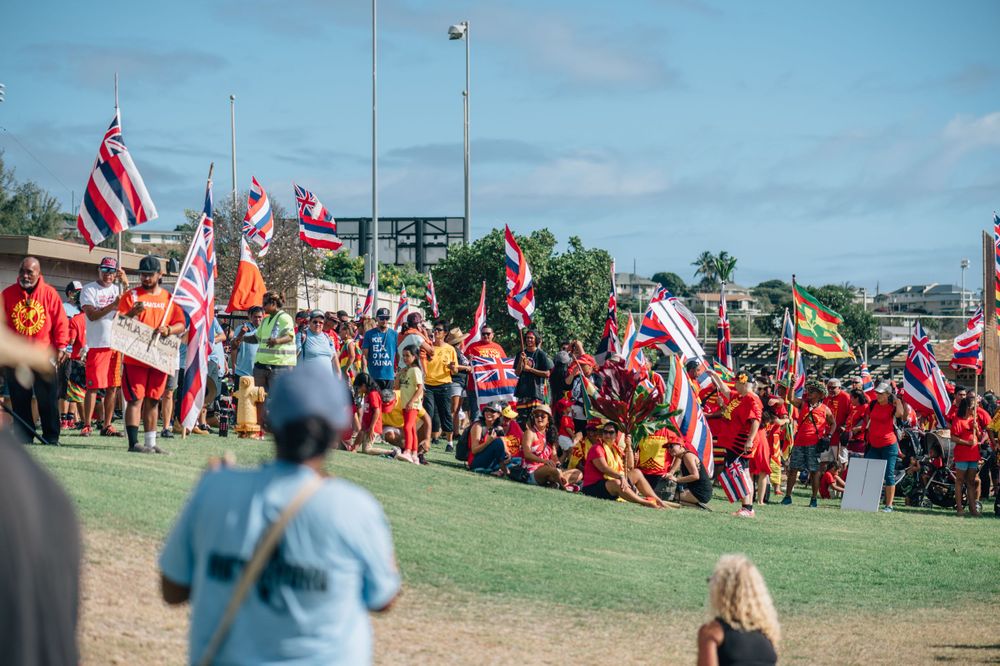 People gathered outdoors with Hawaiian state flags against a blue sky background.