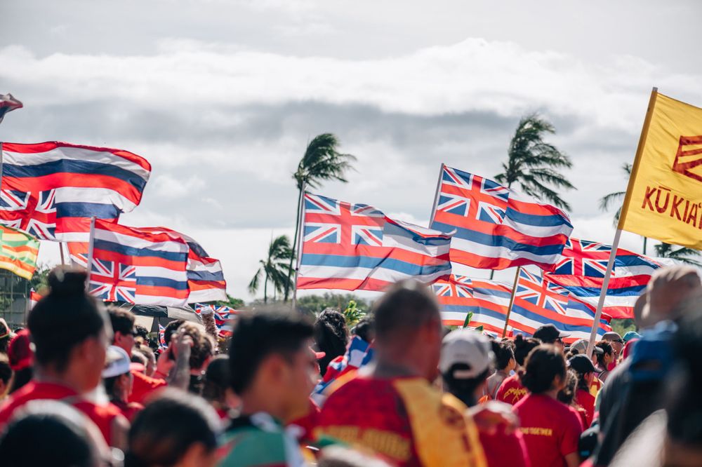Hawaiian state flags wave above a large crowd gathering under cloudy skies.