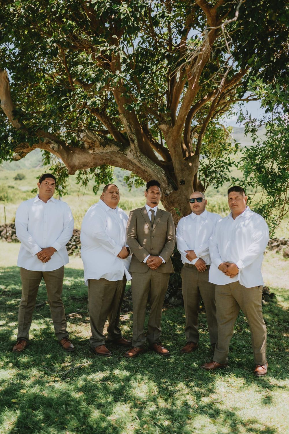 Wedding party members in white shirts and khaki pants pose casually under a shady tree.