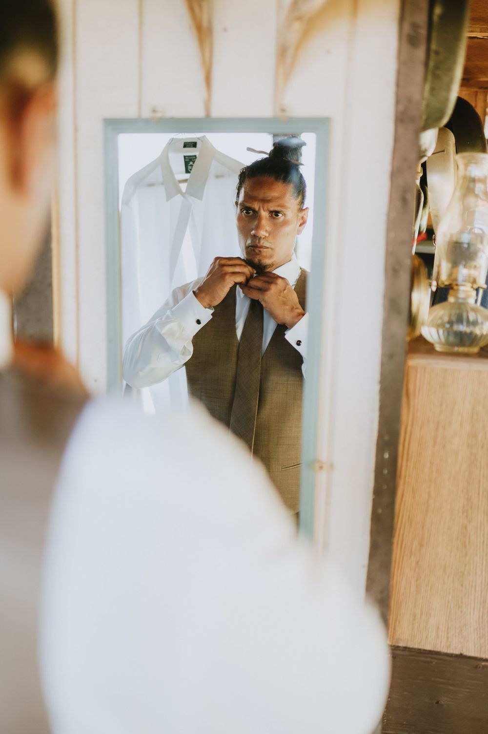 Groom adjusts bowtie while looking in mirror during wedding preparations.