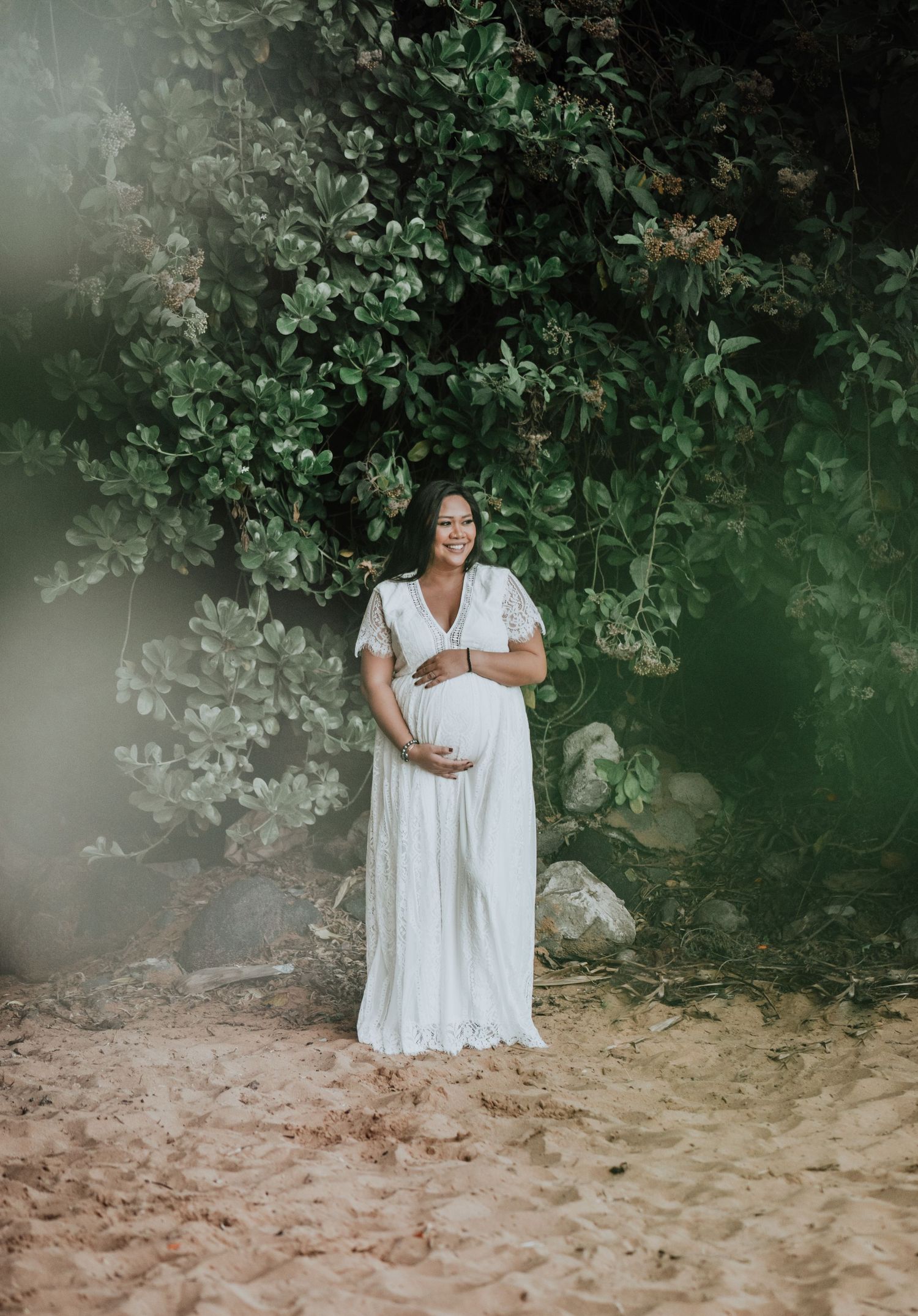 White lace dress flows beautifully against a backdrop of green foliage on a sandy beach.