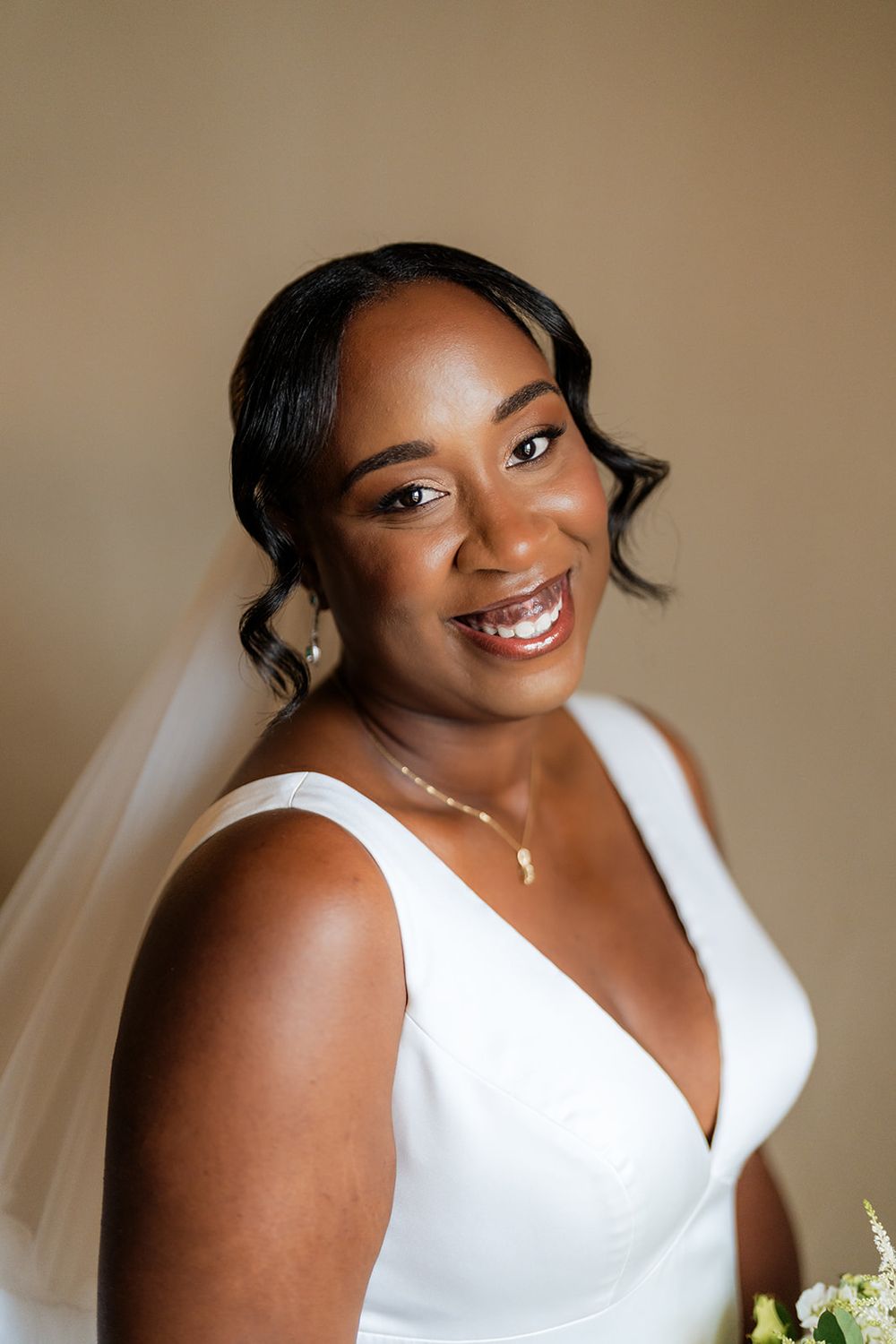 A radiant bride wearing a classic white V-neck wedding dress poses with a joyful expression against a neutral background.