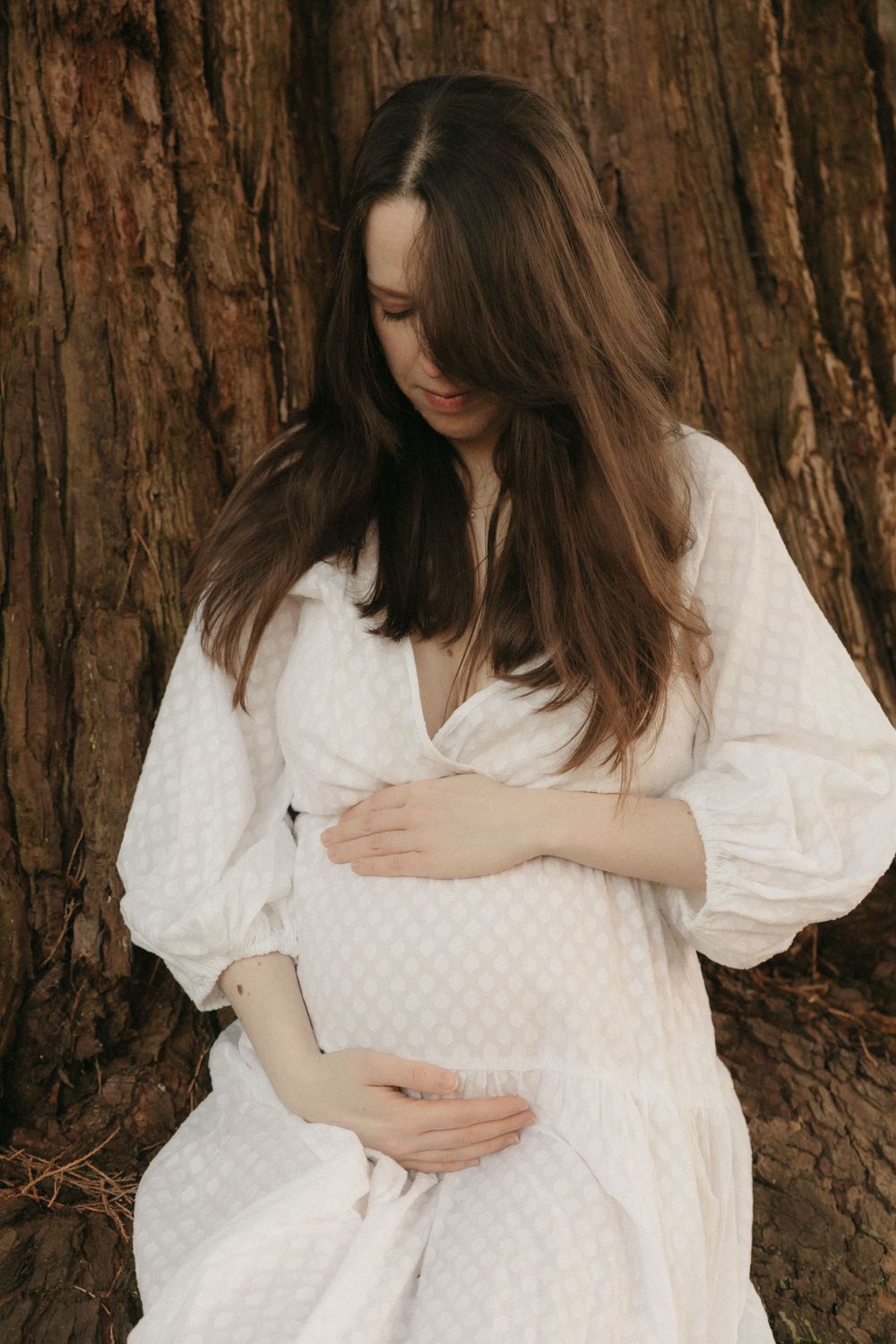 Person in white dress embracing their pregnant belly against rustic tree bark backdrop.