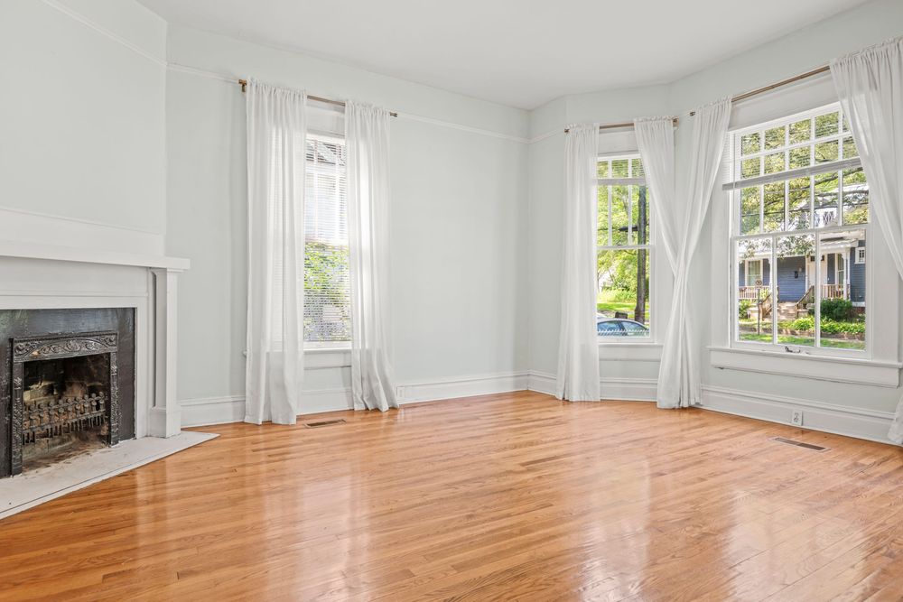 Empty bright room with hardwood floors, white walls, fireplace, and large windows with flowing white curtains.