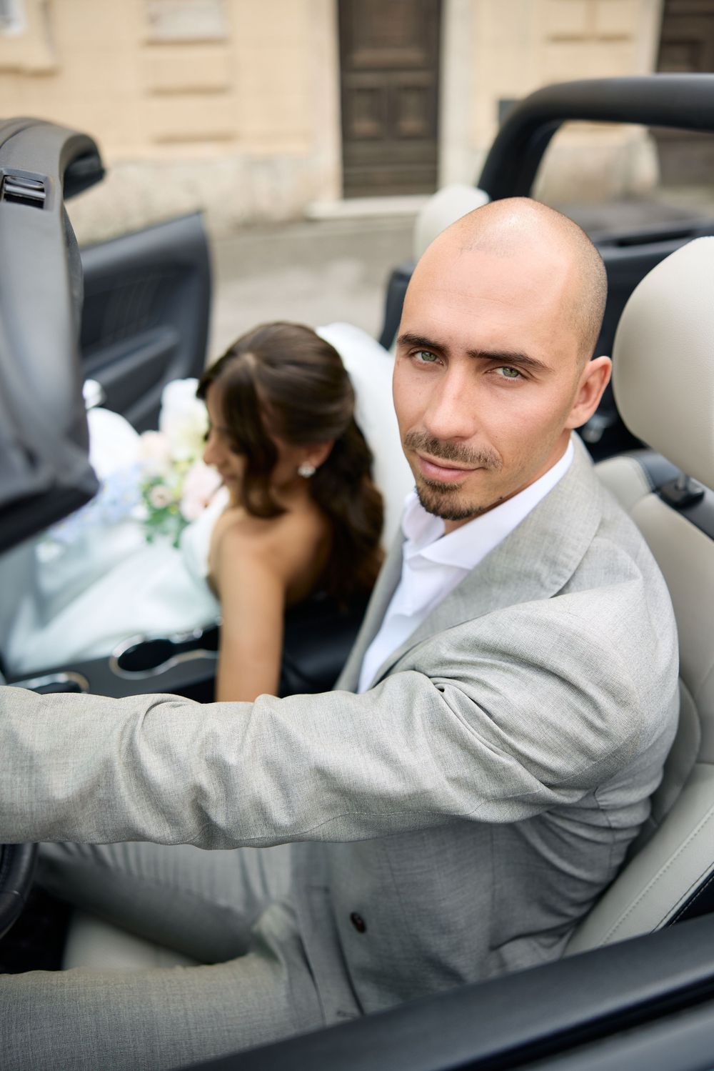 A person in a gray suit sits in a luxury convertible car with black leather interior.