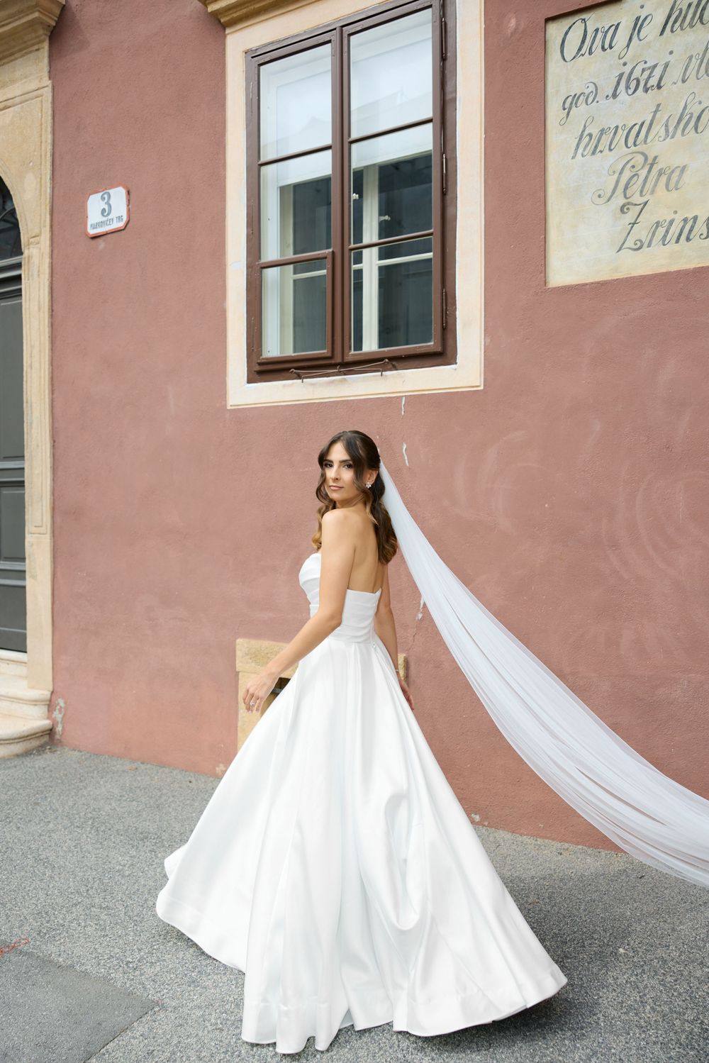 Bride in flowing white dress twirling against pink stucco building wall.