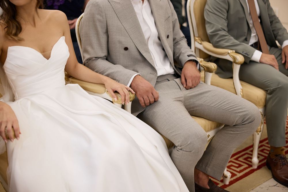 Wedding party members in light gray suits and white dresses sit together on wooden chairs at an outdoor ceremony.