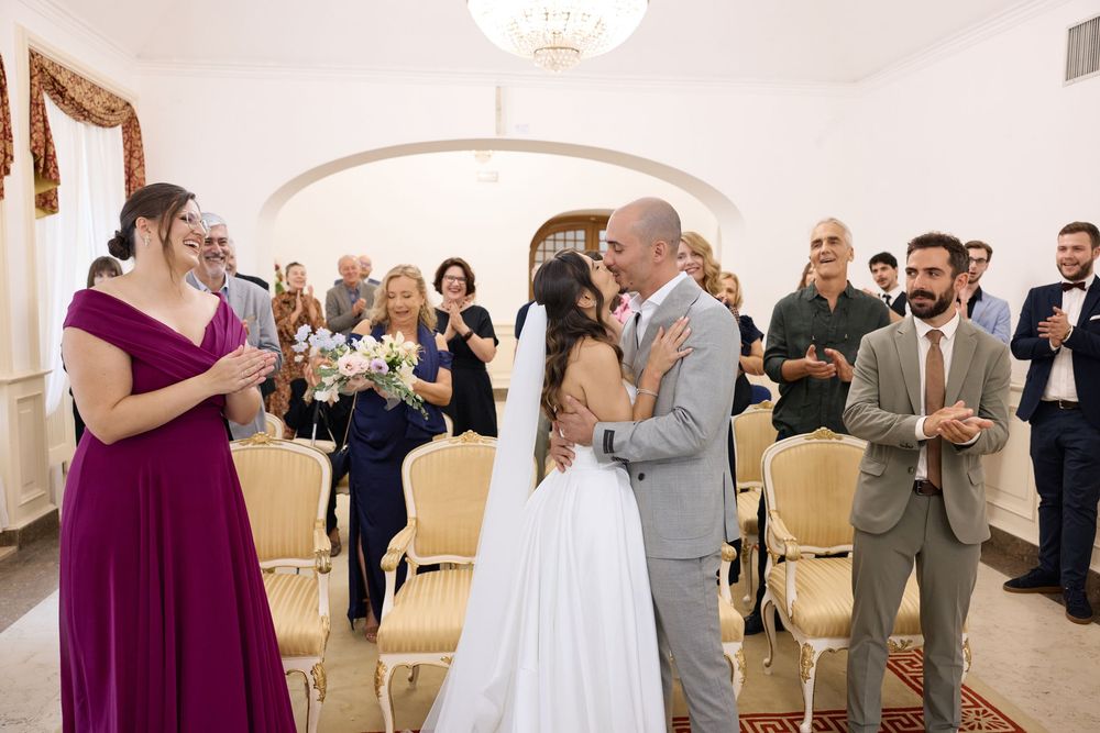 Wedding ceremony guests gathered in an indoor venue with white walls and arched doorways.