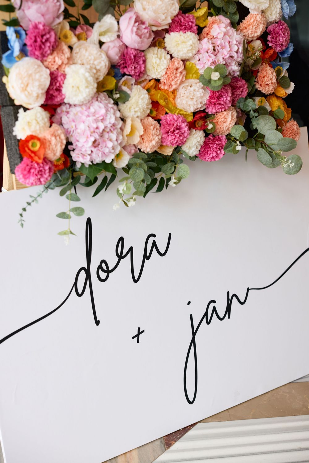 Elegant wedding sign with names 'dora + jan' decorated with pink and peach floral arrangements.