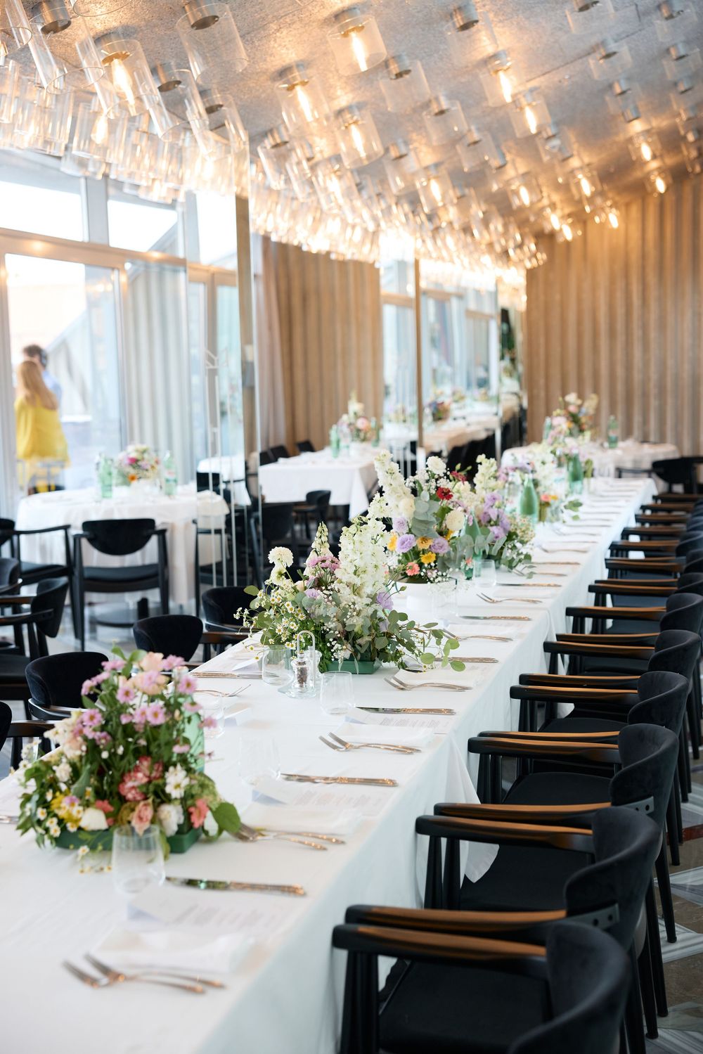 Elegant wedding reception setup with crystal chandeliers, black chairs, and pink floral centerpieces.