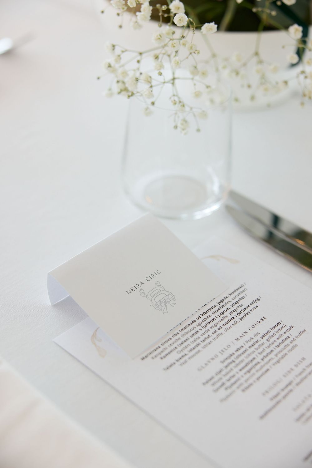 An elegant white wedding menu card displayed on a table with baby's breath flowers and glassware.