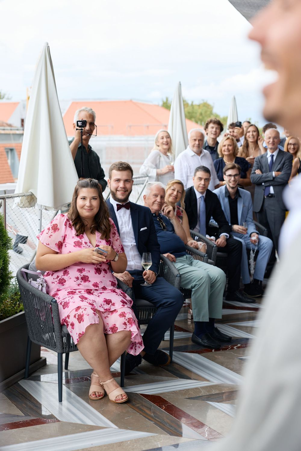 Wedding guests gathered on an outdoor terrace with umbrellas and city rooftops in the background.