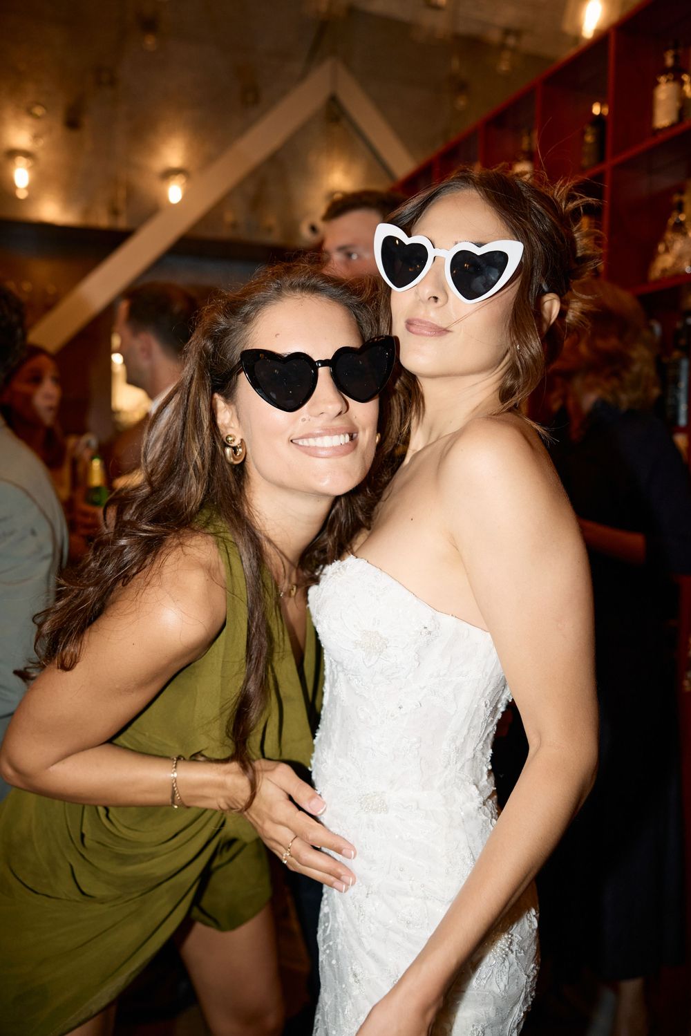 Two friends wearing heart-shaped and black sunglasses pose together at a wedding celebration.