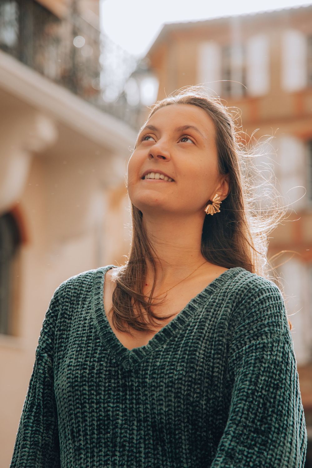 A smiling young woman in a teal knit sweater looks upward on a sunny European street.