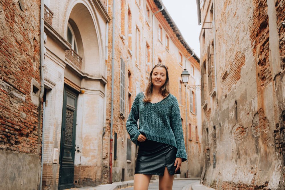 A smiling young woman in a teal sweater and black skirt walks through a narrow European brick alleyway.
