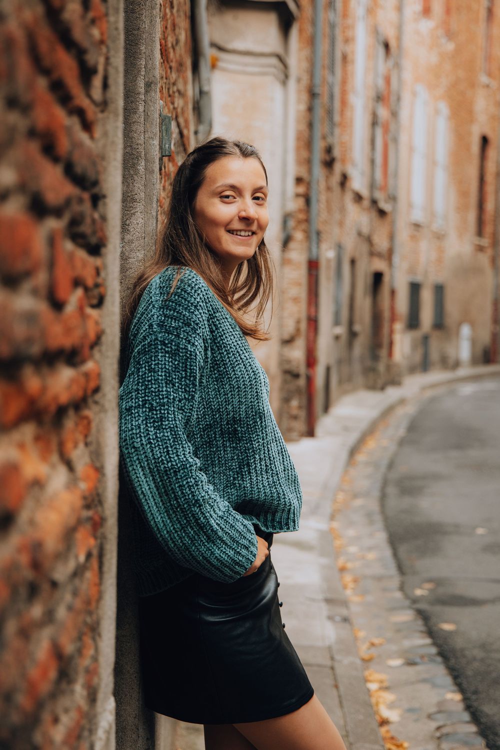 Woman in teal knit sweater and black leather skirt leaning against brick wall in narrow European alley.