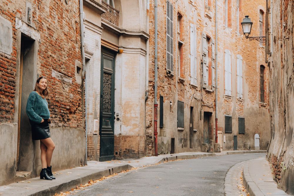 Young woman in teal sweater and black shorts standing in historic European cobblestone alleyway.