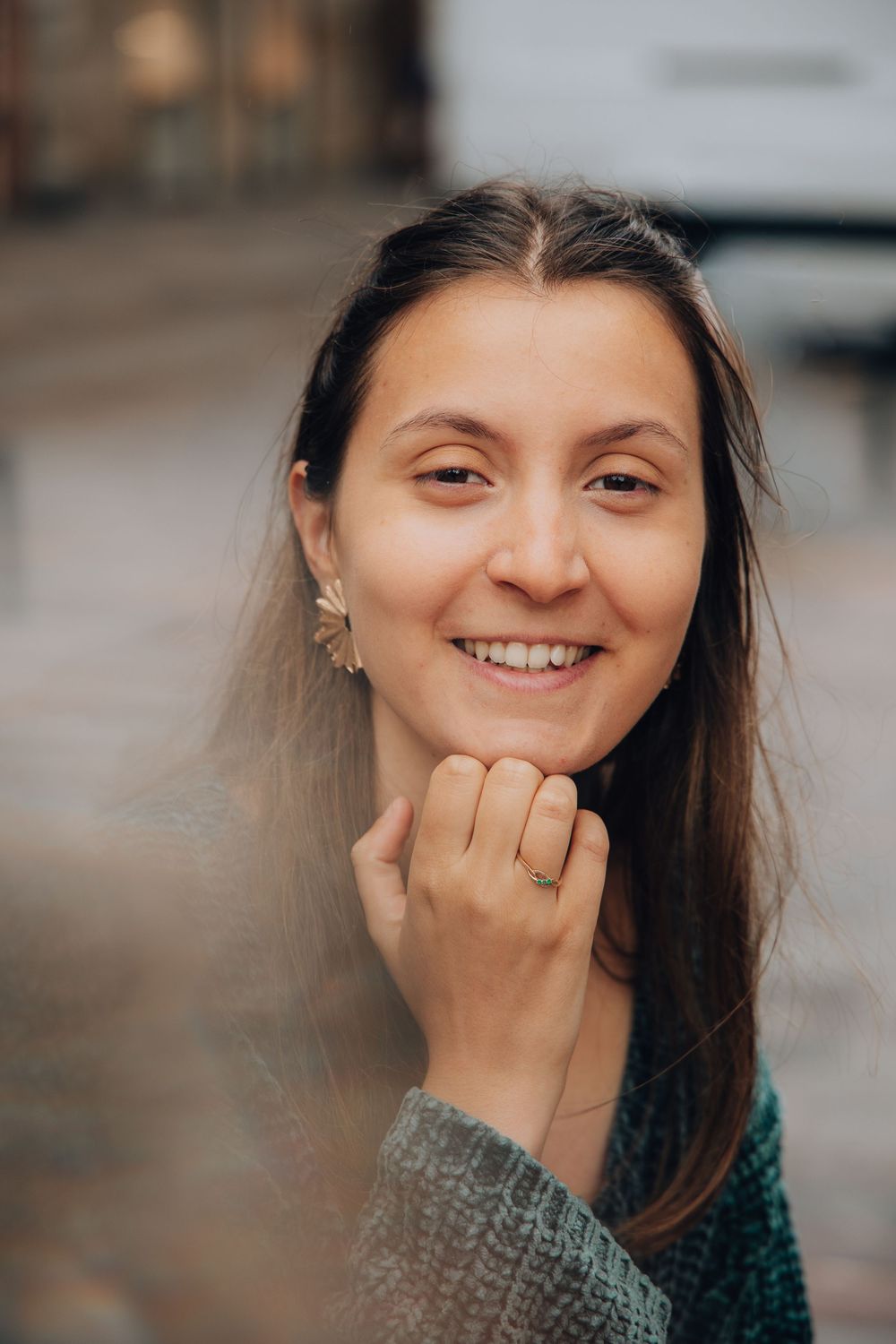 Smiling young woman with dark hair, gold earring and ring, posing with hand near chin outdoors.